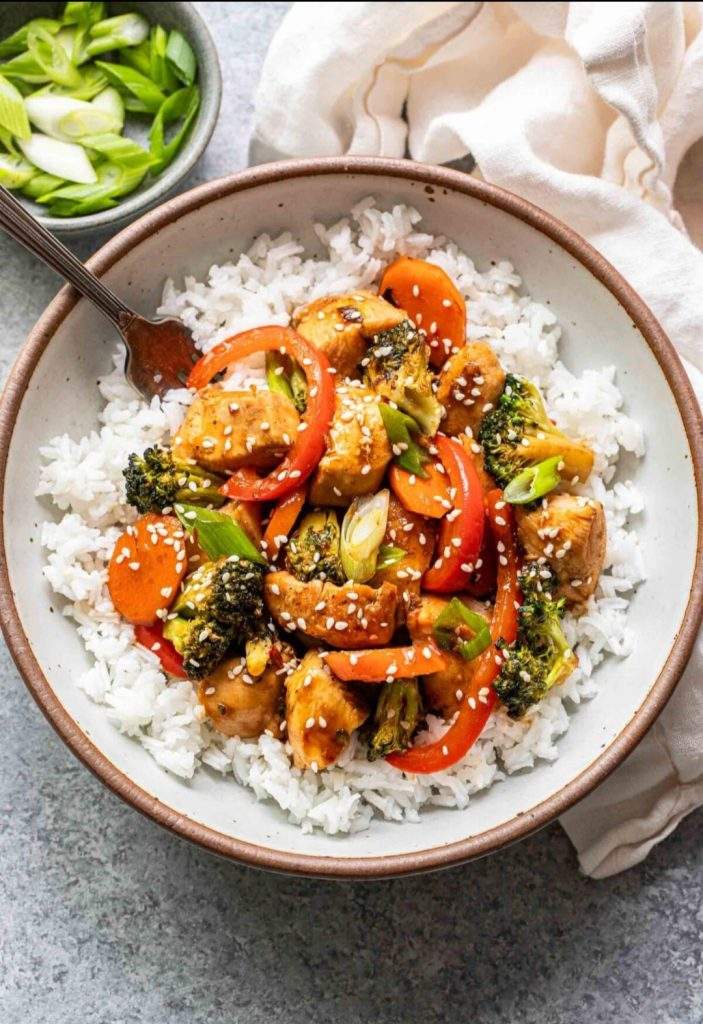 Colorful chicken veggie stir-fry with rice served over white rice in a bowl, featuring bell peppers, broccoli, carrots, and a sprinkle of sesame seeds on top.