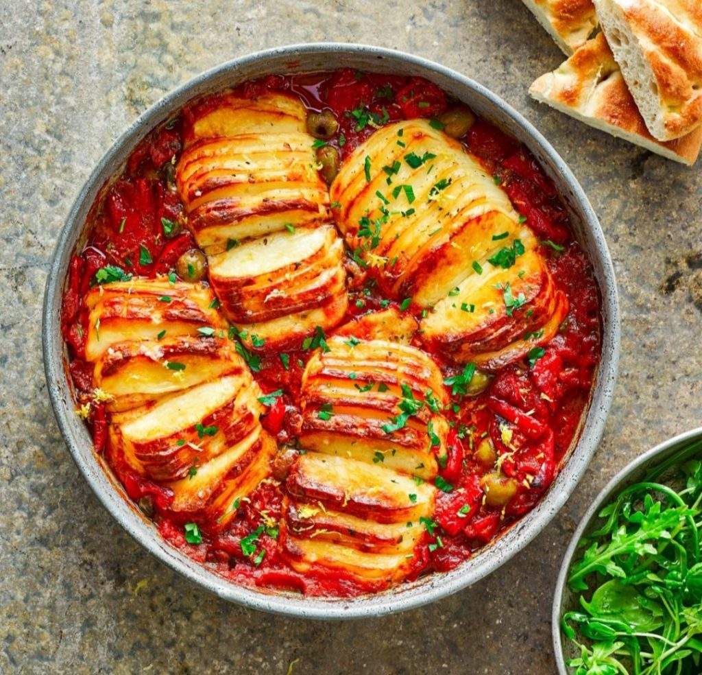 Golden baked hasselback halloumi in a tray with tomato, olives, and capers – served with flatbreads and fresh salad.